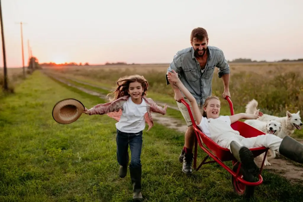 Family playing in a sunny field with a wheelbarrow and their dog