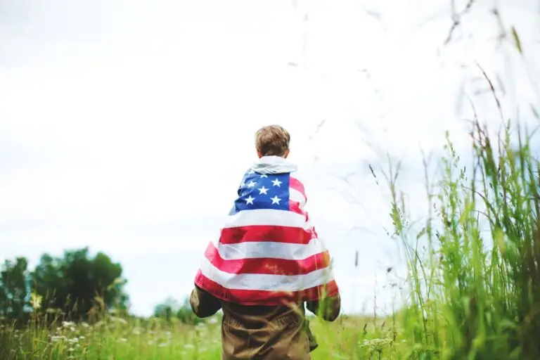 Child wrapped in an American flag riding on a parent's shoulders in a meadow