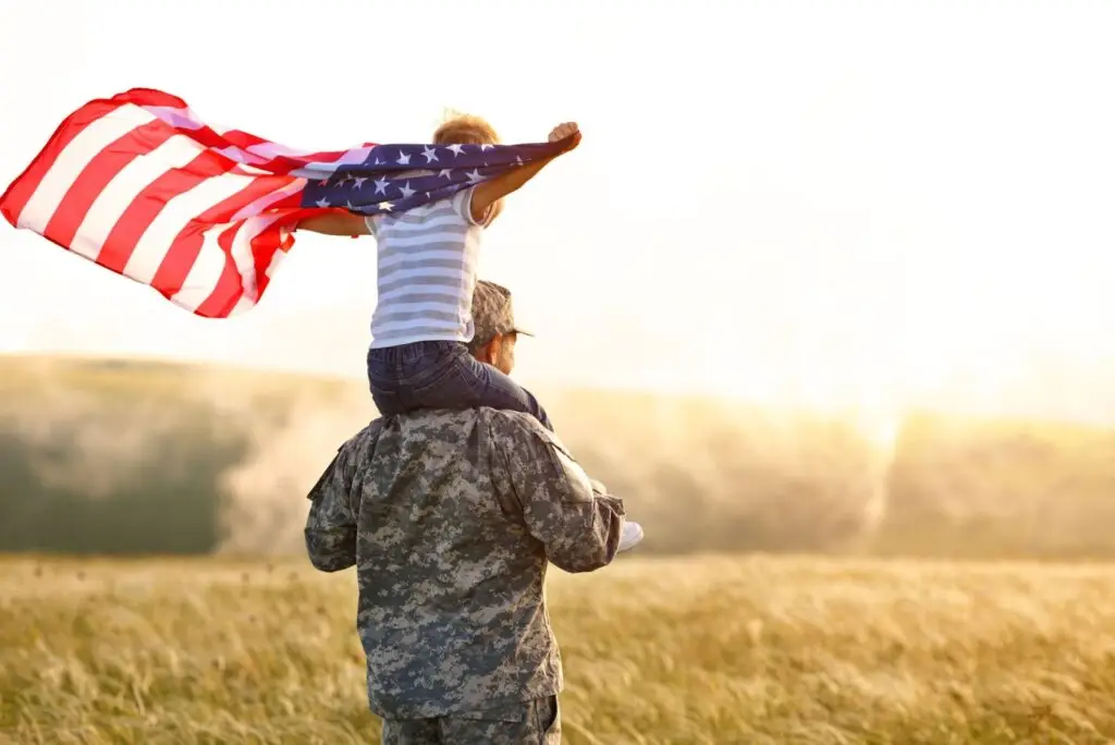 Service member in uniform carrying a child who is waving an American flag
