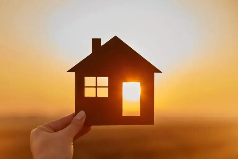 Hand holding a house shaped cutout up to a glowing orange sunset