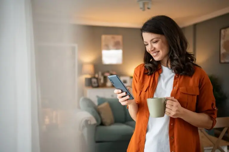Woman in an orange shirt smiling at her phone and holding a coffee mug