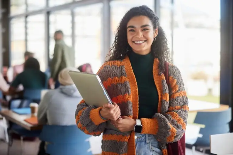 Smiling student in a striped cardigan holding a laptop in a busy cafe