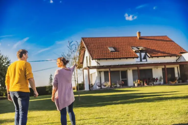 Couple walking across a sunny lawn toward a white house with a red roof