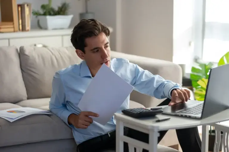 Man in a blue shirt reviewing a document beside his laptop at home